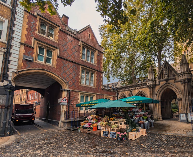 A street scene featuring a flower stall set up under a green umbrella on cobblestone pavement, displaying a vibrant variety of fresh flowers including roses, lilies, and carnations in different colors