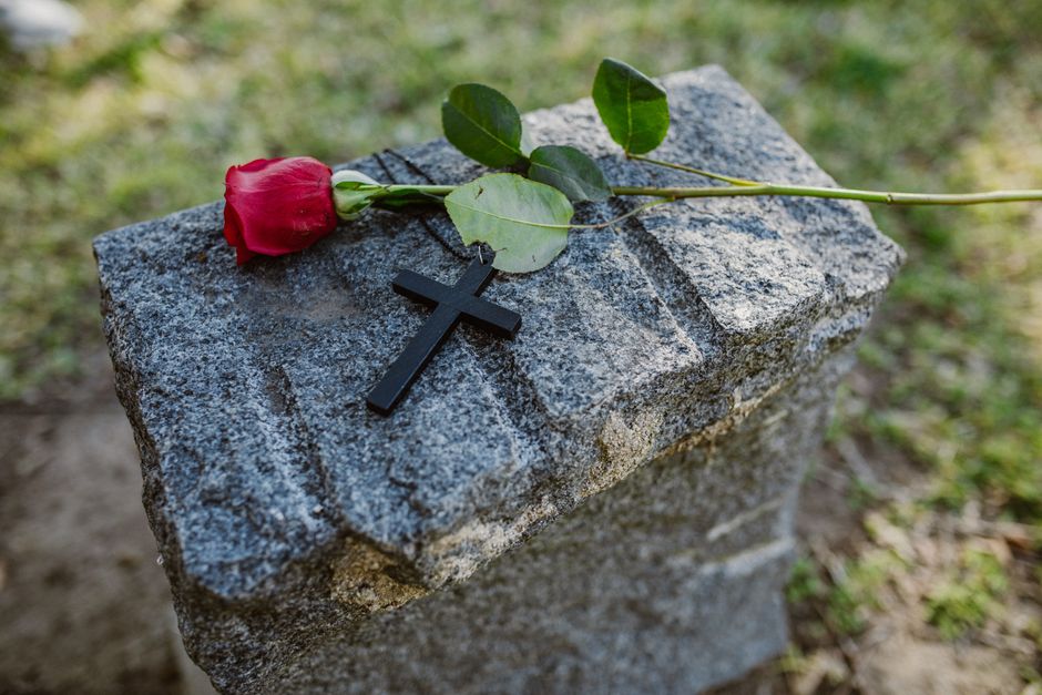 A single deep red rose with lush, velvety petals and fresh green leaves resting on a textured grey granite memorial stone. The rose's long green stem extends across the stone, while a small black wood