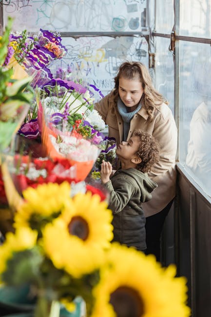 A woman and a young girl are inside a flower shop, browsing a vibrant display of fresh floral arrangements. The woman has wavy light brown hair and is wearing a beige coat, while the girl has curly da