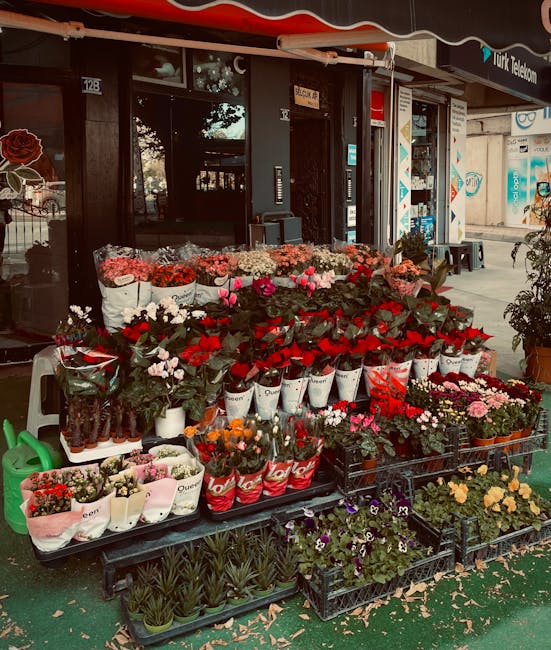 An outdoor display of fresh flowers in front of a florist shop, with a variety of vibrant blooms arranged in white and red buckets and black crates on a raised black table. The arrangement includes pi